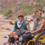 Two women in wheelchairs smiling in the Grand Canyon along the Colorado River